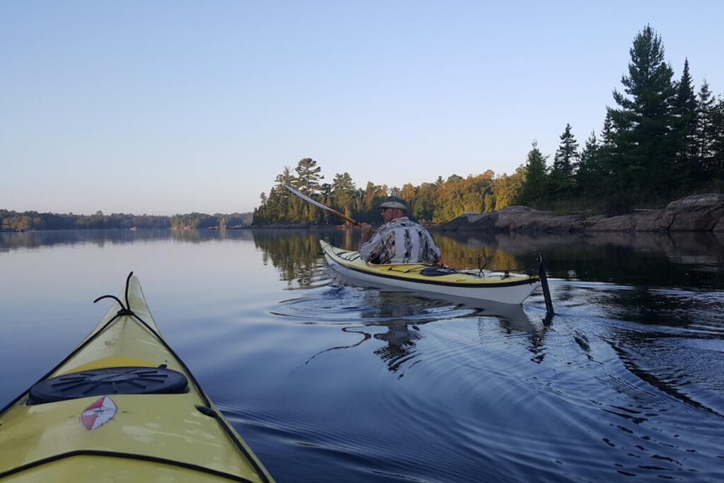 A photo taken from the seat of a yellow and black kayak on a lake, with trees and rock surrounding the shoreline. A person wearing a shirt decorated with kayaks paddles an identical yellow and black kayak in the center of the photo.