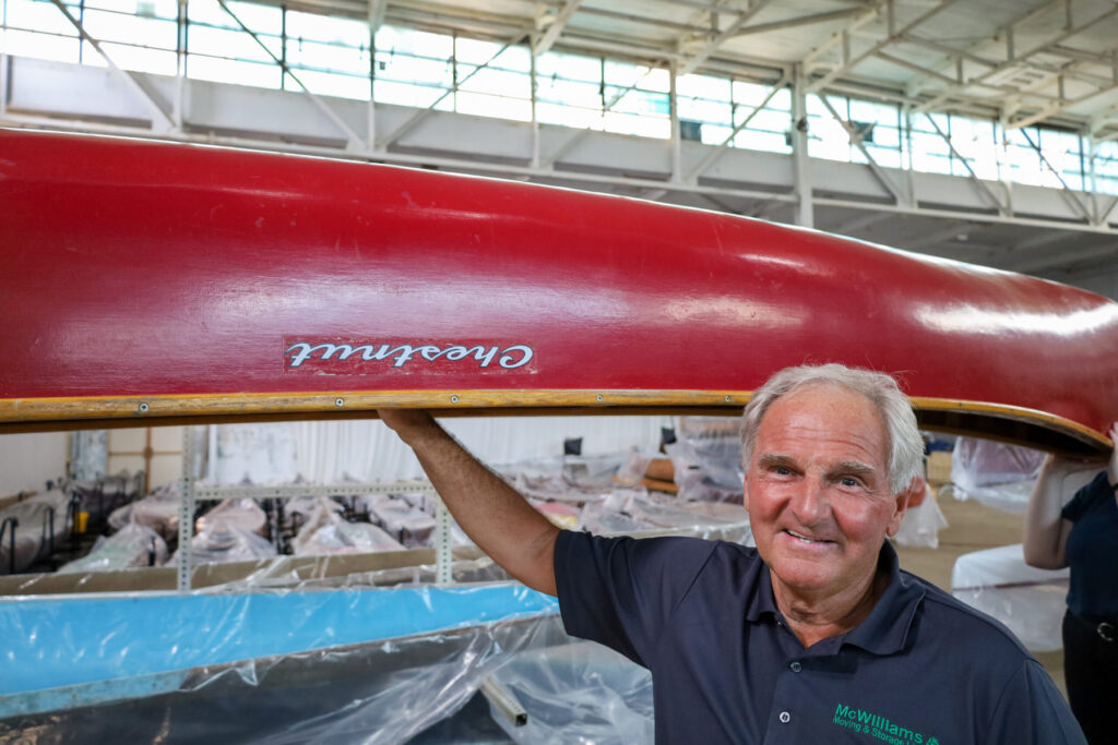 Dan McWilliams, of McWilliams Moving & Storage Ltd, stands smiling in a blue branded polo shirt with his hand on an overturned red Chestnut wooden canoe in the Canadian Canoe Museum's old Collection Centre at 910 Monaghan Road. Canoes wrapped in plastic can be seen in the background.