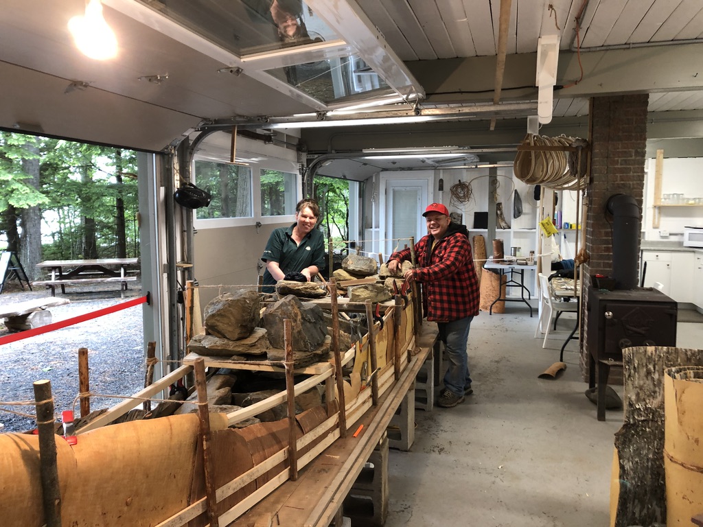 Two people pose at the end of a partially constructed birchbark canoe