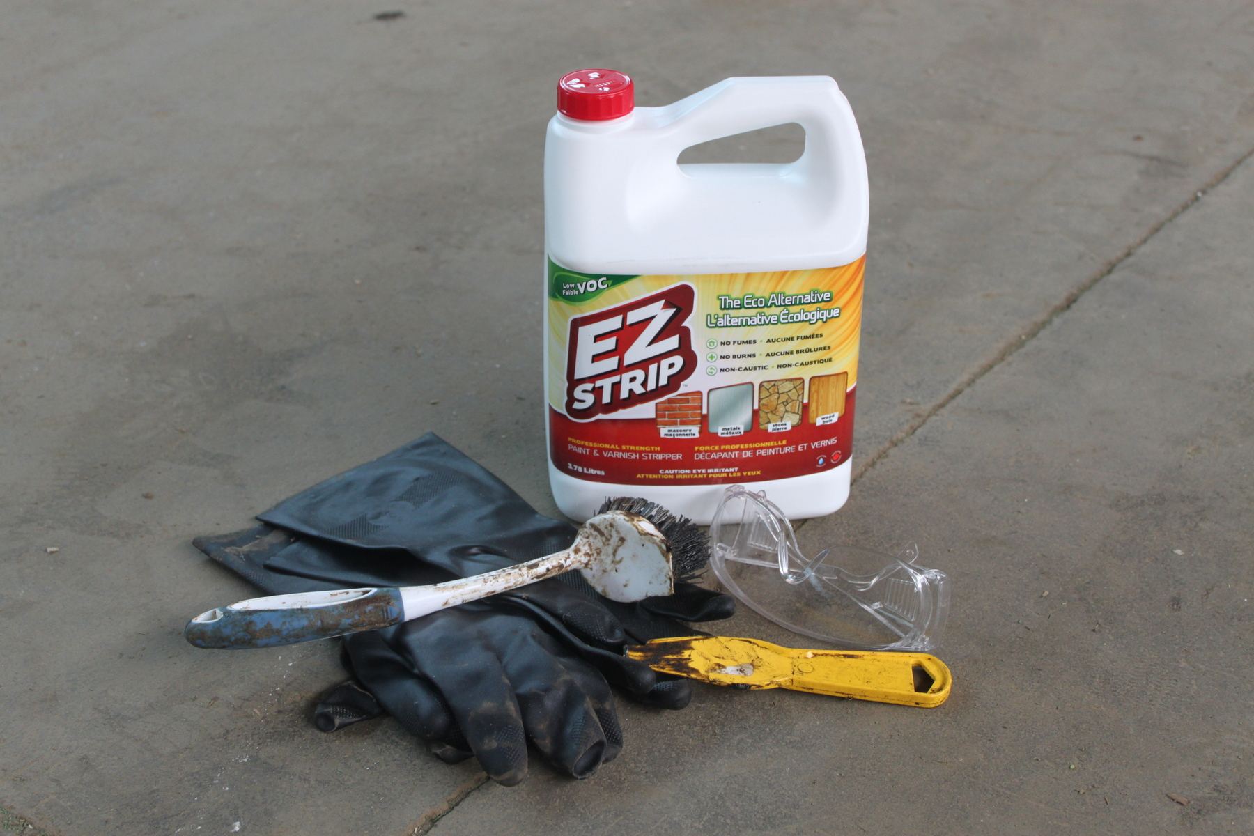 Various restoration materials displayed on the floor, including a white bottle of varnish, black rubber gloves, a strub brush, goggles and a yellow scraper.