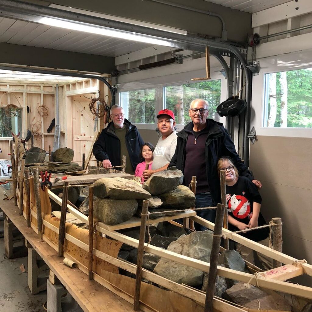 Group of people pose while standing in front of a canoe in a building shop