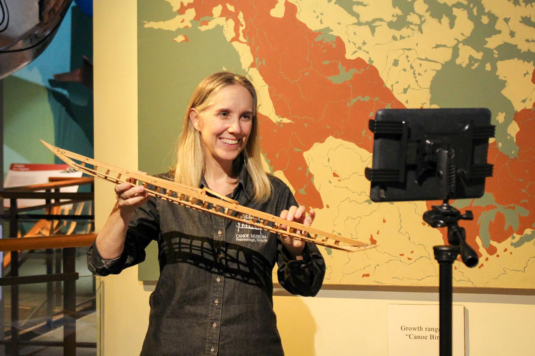 A Canadian Canoe Museum educational staff member holds a model of a kayak frame while smiling and standing in front of an iPad on a stand during a virtual field trip.