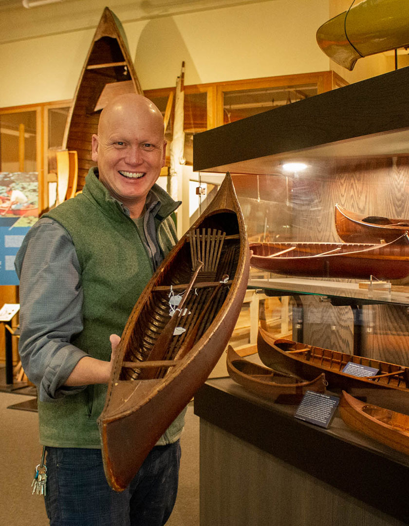 Curator Jeremy Ward stands in front of a case of model canoes, smiling and holding one of the canoes towards the camera.