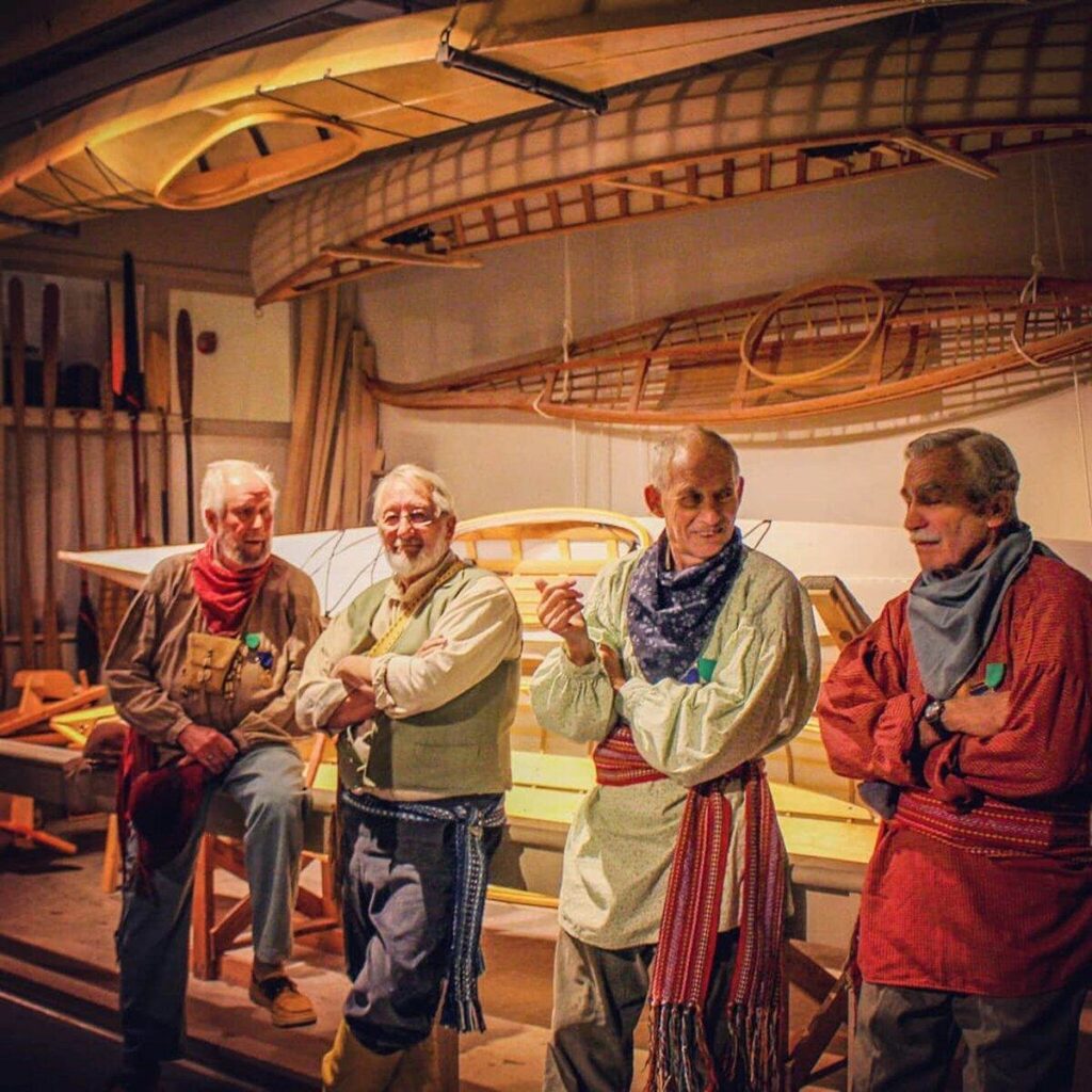 Woodshop volunteers dressed as voyageurs standing together in front of canoe display.