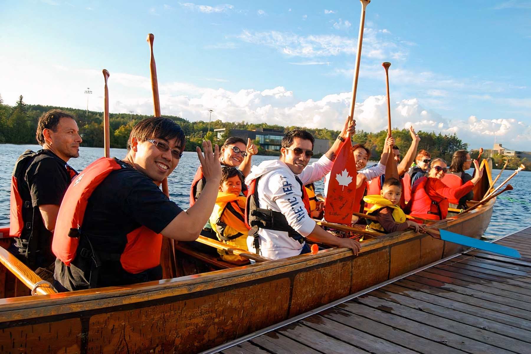 A group of adults and childrens raise their paddles and smile in a Voyageur Canoe about to embark on a paddling tour.