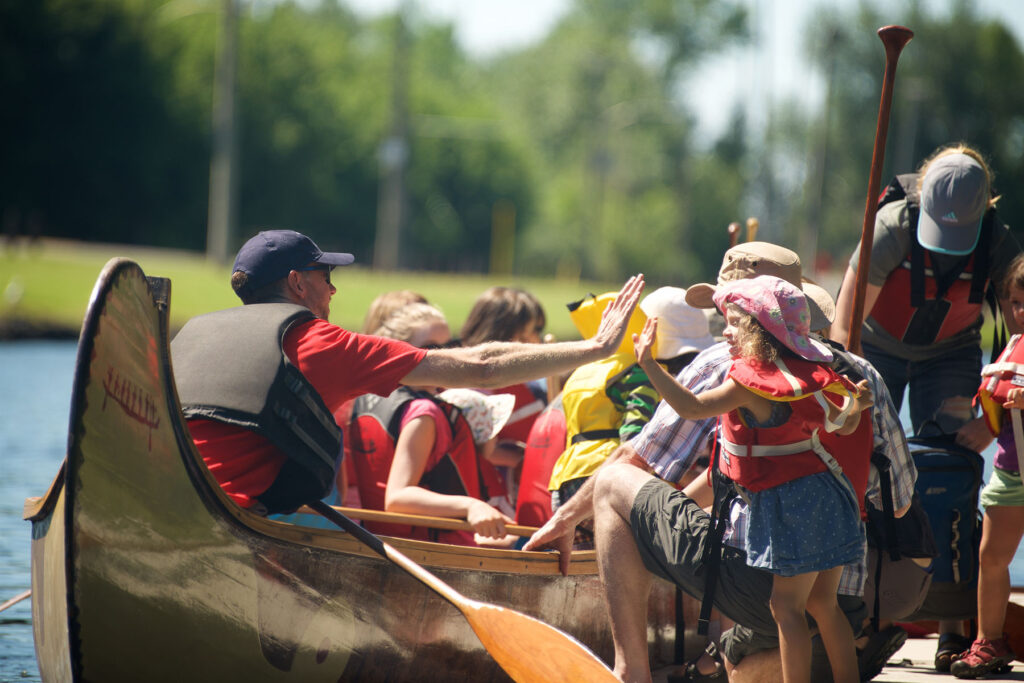 A Canadian Canoe Museum instructor in a voygageur canoe filled with adults and youth high-fives a young girl on the dock.