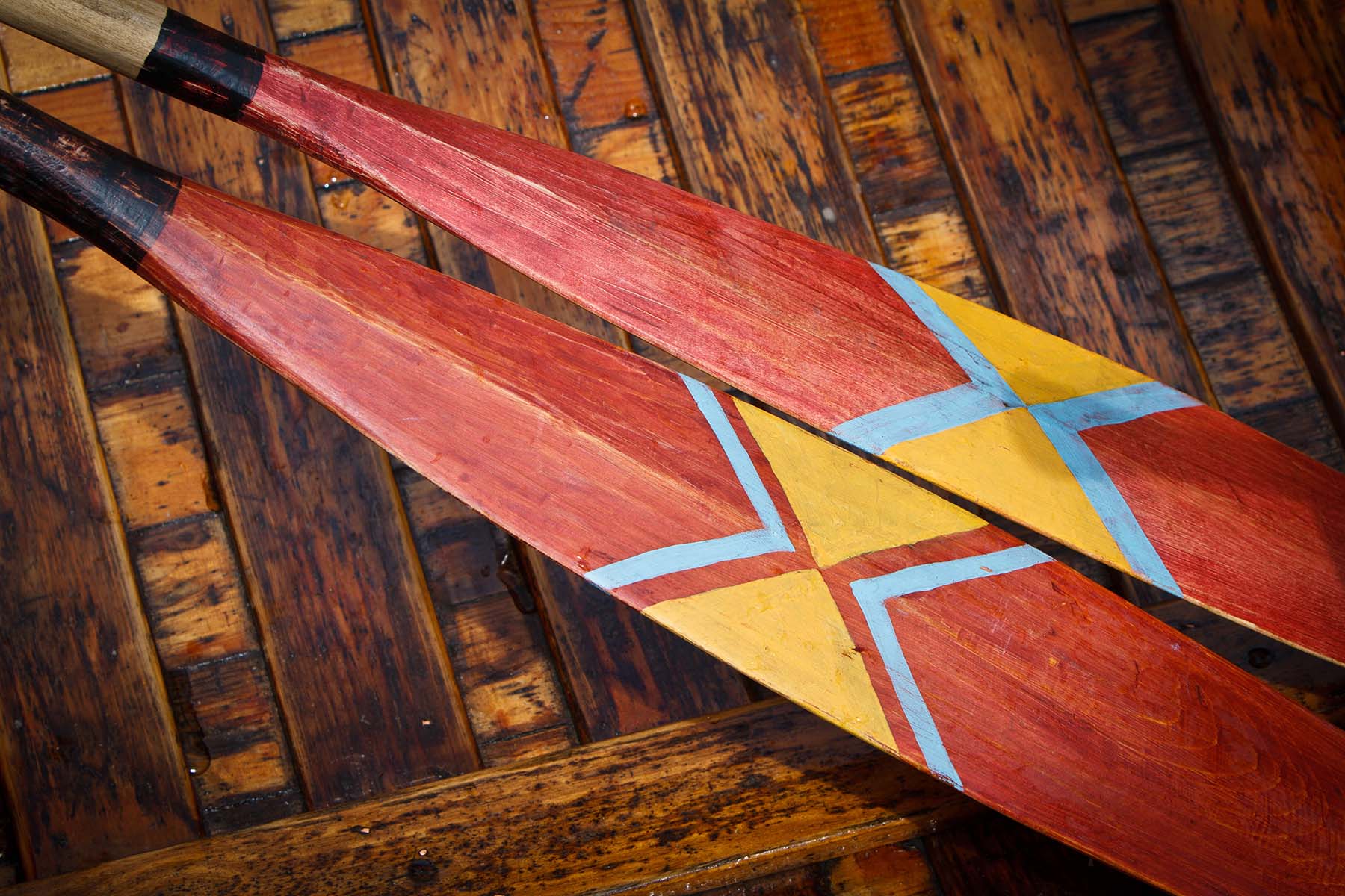 Two voyageur canoe paddles with blue and yellow designs lay side by side against the bottom of a wooden canoe.