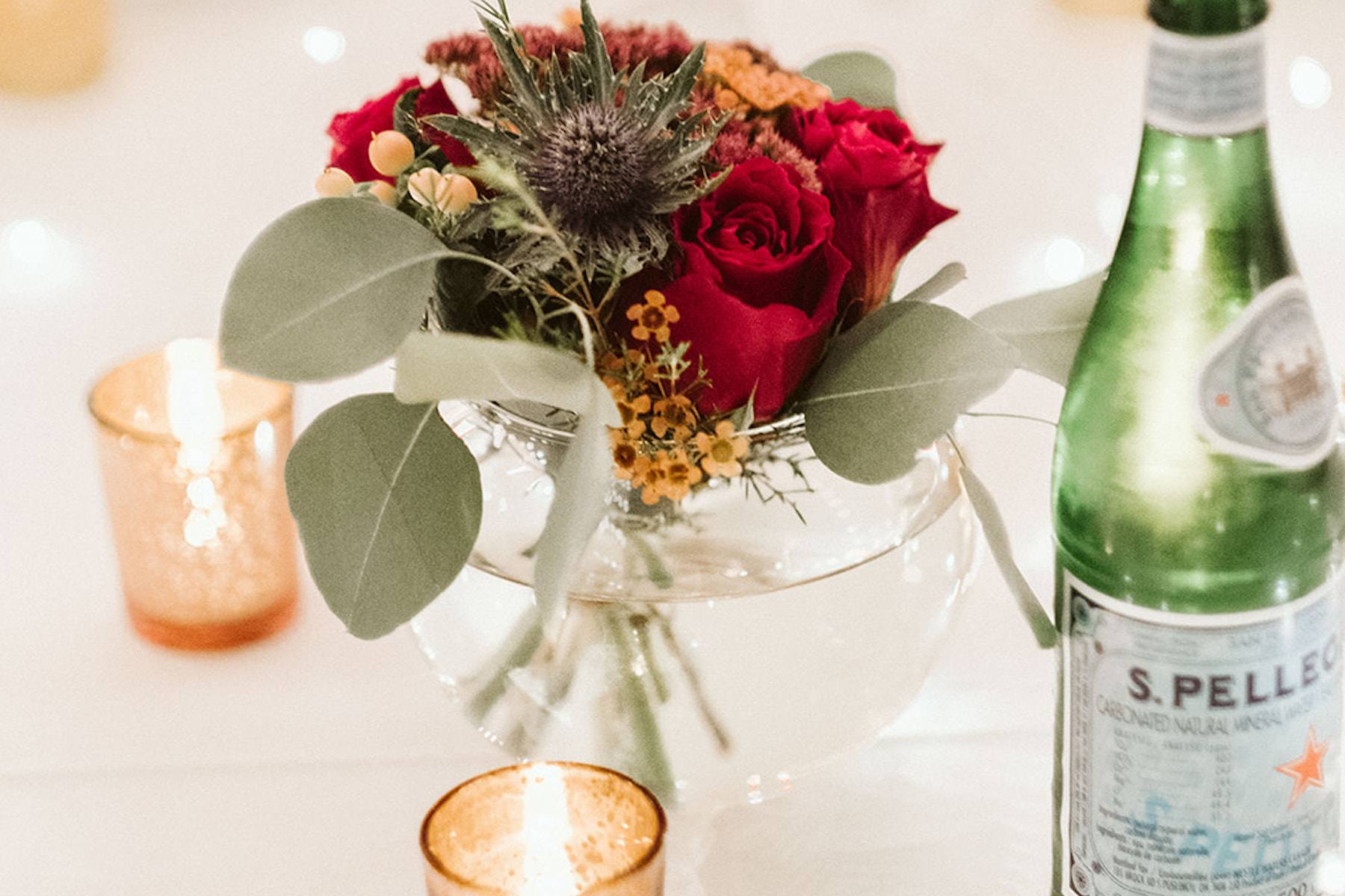 A bouquet of green, red, and yellow flowers sits in the middle of a table, surrounded by candles and a bottle of sparkling water.