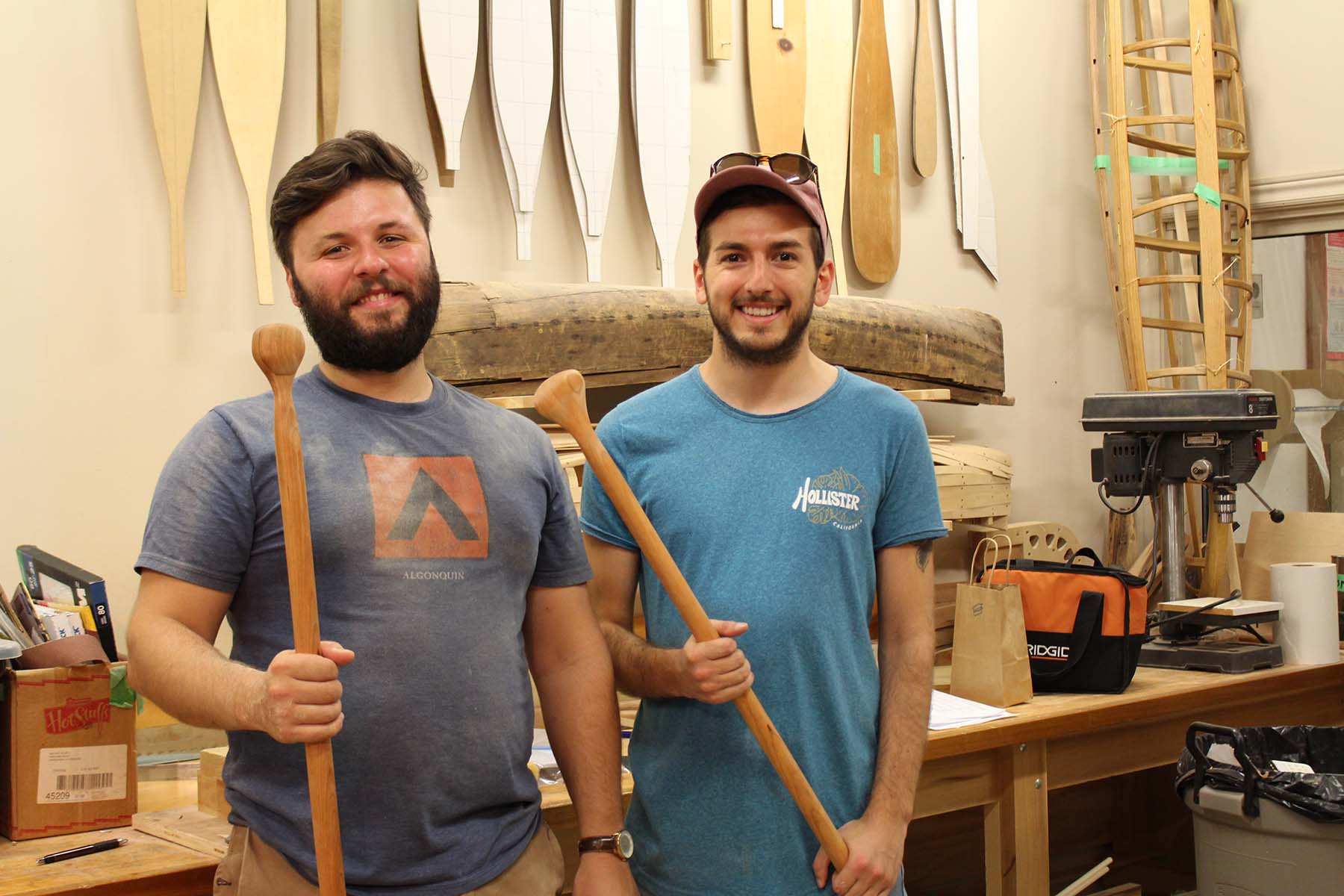 Two men stand smiling in a workshop holding two paddles that they hand-carved themselves during an artisan workshop at The Canadian Canoe Museum.