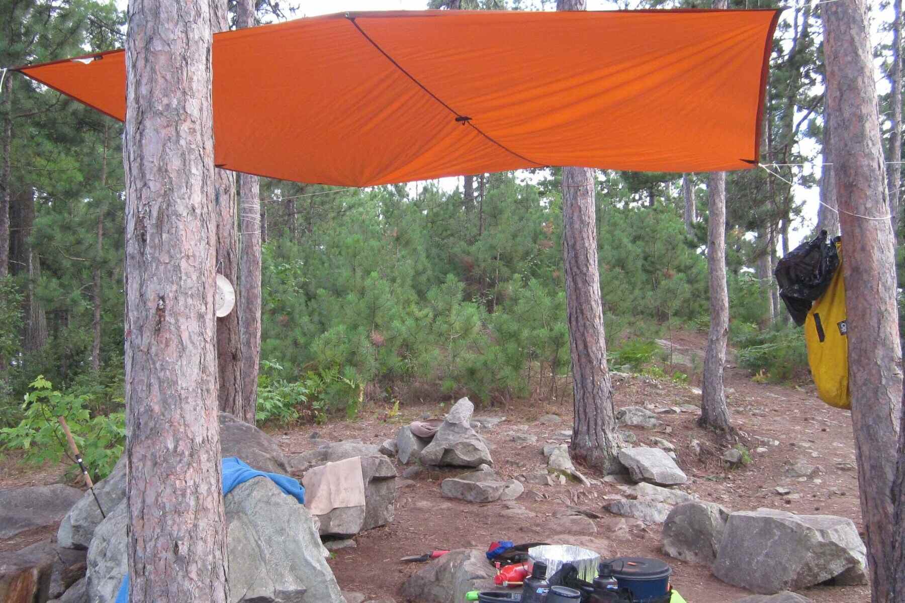 An orange tarp is tied up to provide a cover over a small camp site surrounded by evergreen trees