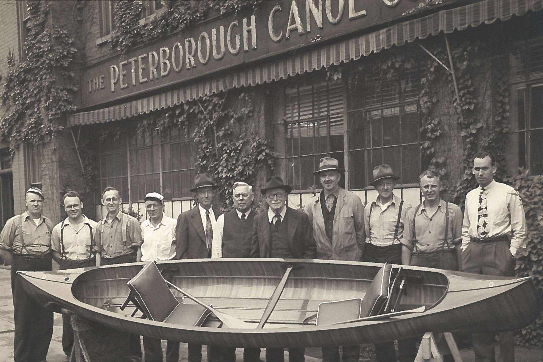 Black and white photo of several men lined up behind a canoe, posing for a photograph.