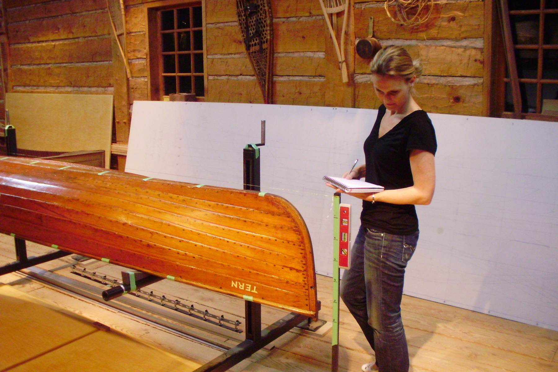 A lapstrake canoe rests upside down as someone stands nearby holding a notebook