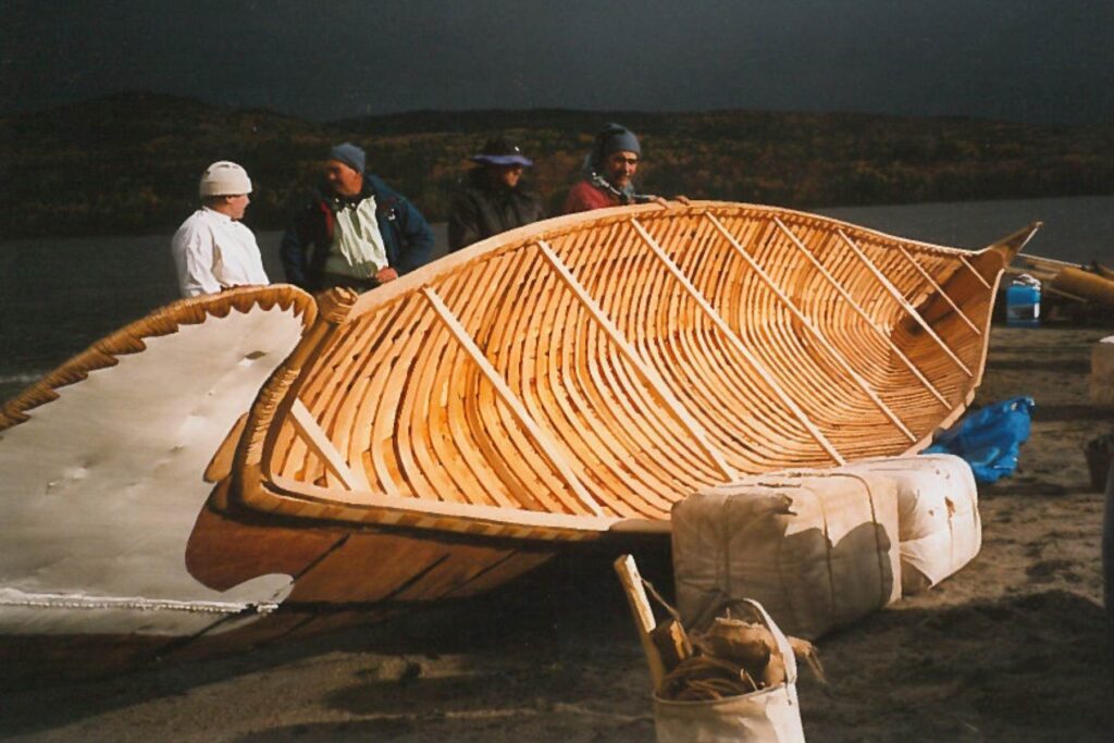 Outside at night, a large birch canoe sits on its side with a few cargo packages nearby.