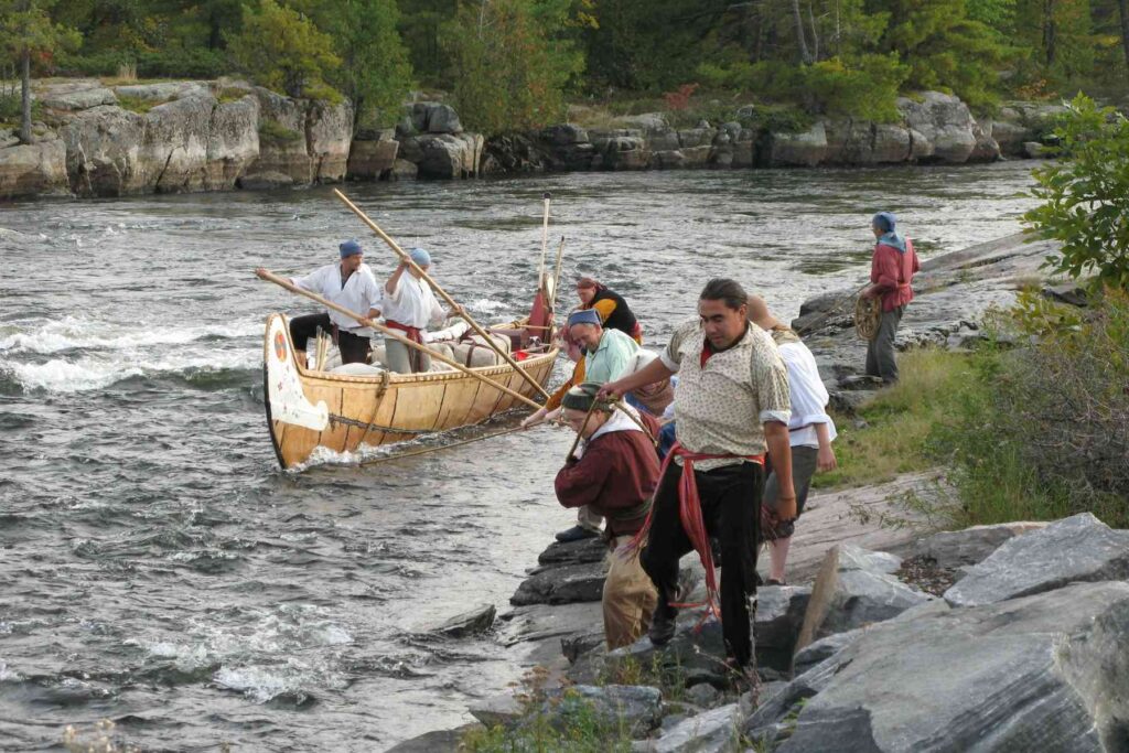 Several people stand along a rocky riverbank while two paddlers navigate a large birch canoe upstream through small rapids.