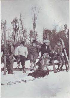 Black and white photograph of people sitting along a fence holding their winter skis